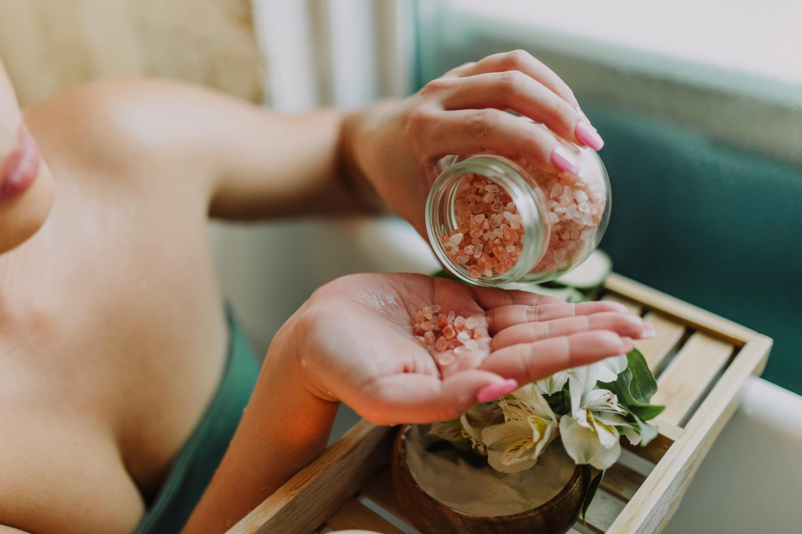 Close-up of hands pouring pink bath salt with flowers in a relaxing spa setting.